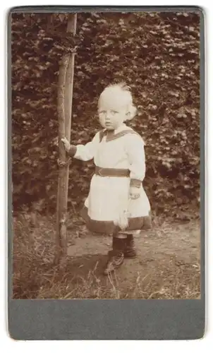 Fotografie unbekannter Fotograf und Ort, blondes süsses Mädchen im Garten am Baum stehend