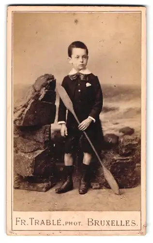 Fotografie Fr. Trable, Bruxelles, Rue de la Madeleien 56a, Portrait kleiner Knabe im Anzug mit Paddel in der Hand