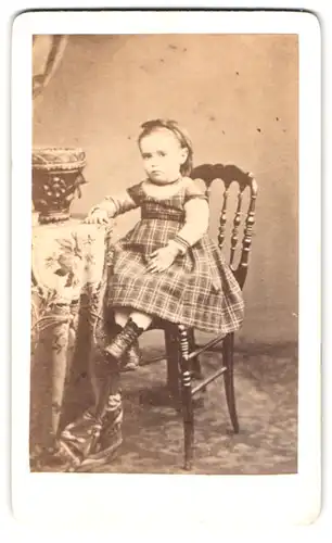 Fotografie Fotograf und Ort unbekannt, Portrait Mädchen im karierten Kleid mit Haarreif sitzend auf einem Stuhl