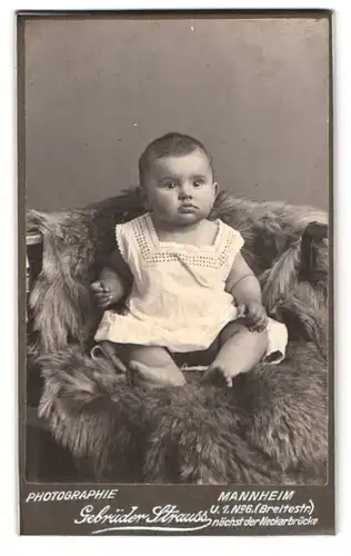 Fotografie Gebr. Strauss, Mannheim, U. 1. No. 6, Portrait Kleinkind im Kleidchen auf einem Fell sitzend