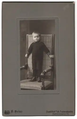 Fotografie B. Heinz, Frankfurt a. M., Brückenstr. 54, Portrait kleiner Junge im schwazen Overall steht auf einem Stuhl