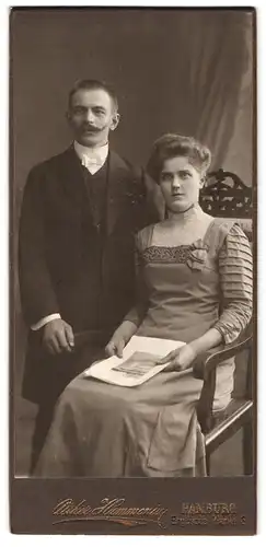 Fotografie Atelier Hammonia, Hamburg, Englische Planke 6, Herr mit langem Mantel steht neben sitzender Dame in Kleid