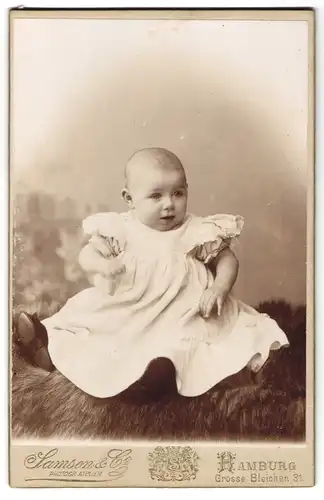 Fotografie Samson & Co., Hamburg, Grosse Bleichen 31, niedliches Baby im Taufkleid auf Felldecke sitzend