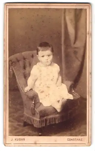 Fotografie J. Kuban, Constanz, Husenstr. 30, Portrait Kleinkind im weissen Kleid sitzt im Kindersessel
