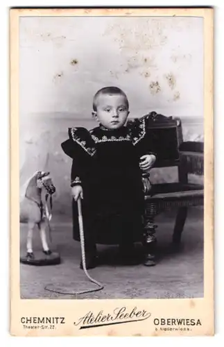 Fotografie Seeber, Chemnitz, Theater-Str. 22, Portrait kleines Kind im schwarzen Kleid mit Peitsche und Spielzeugpferd
