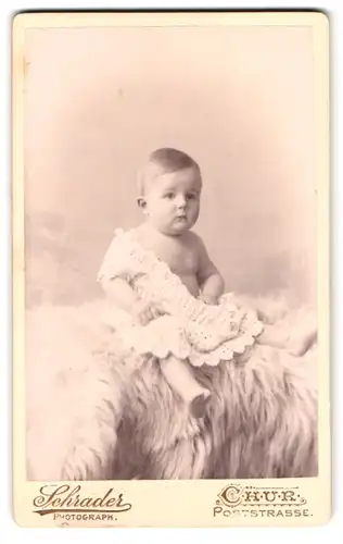 Fotografie Schrader, Chur, Poststr., Portrait Kleinkind im weissen Kleid auf einem Fell sitzend