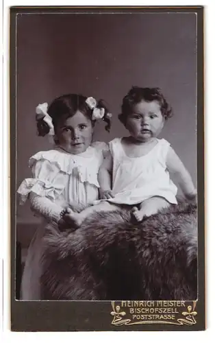 Fotografie Heinrich Meister, Bischofszell, Portrait zwei Kinder in Kleidern auf einem Fell, Haarschleife