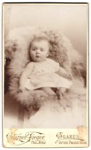Fotografie Glarner Fieger, Glarus, Untere Pressistrasse, Niedliches Baby im weissen Kleid sitzt auf weichem Fell