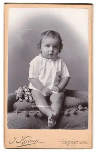 Fotografie J. Kuban, Konstanz, Hieronymusgasse, Portrait Kleinkind im weissen Leibchen mit Puppen an der Seite