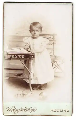 Fotografie Weingartshofer, Mödling, Hauptstr. 79, Portrait kleines Mädchen im weissen Kleid steht am Tisch