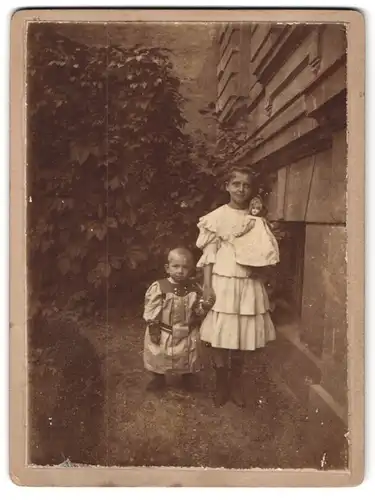 Fotografie Fotograf und Ort unbekannt, Portrait zwei Geschwister in Kleidern mit Puppe in der Hand
