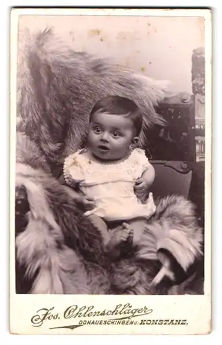 Fotografie Jos. Ohlenschläger, Konstanz, Hussenstr. 15, Portrait bezauberndes Kleinkind im Hemdchen auf Fell sitzend