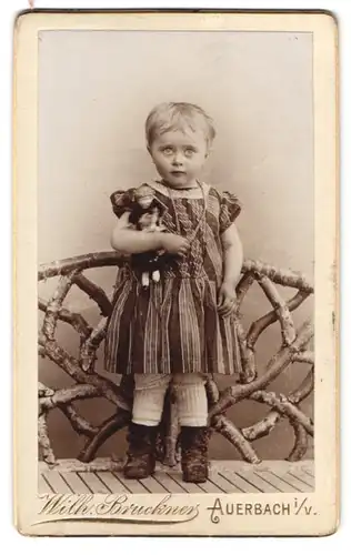 Fotografie Wilh. Bruckner, Auerbach i. V., Portrait kleines Mädchen im gestreiften Kleid mit Puppe in der Hand