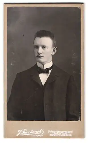 Fotografie J. Paczykowski, Ort unbekannt, Portrait junger Mann im schwarzen Anzug mit Fliege, Bürstenhaarschnitt
