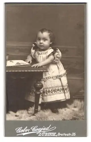 Fotografie Gebr. Coujad, Lübeck, Breitestr. 35, Portrait kleines Kind im Kleid hällt sich am Tisch fest