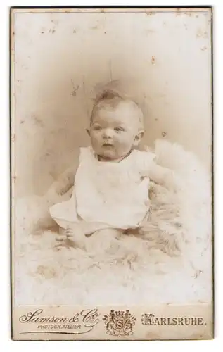 Fotografie Samson & Co., Karlsruhe, Portrait süsses Baby im weissen Kleidchen auf Fell sitzend