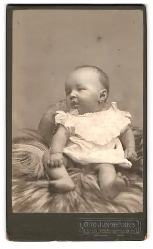 Fotografie Otto Junghans, Leipzig-Gohlis, Georgstr. 22, Portrait süsses Baby im weissen Kleidchen auf Fell sitzend
