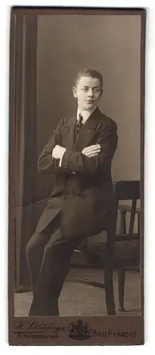 Fotografie H. Schlitzberger, Bad Pyrmont, Portrait junger Knabe im schwarzen anzug mit Schlips und verschrenkten Armen