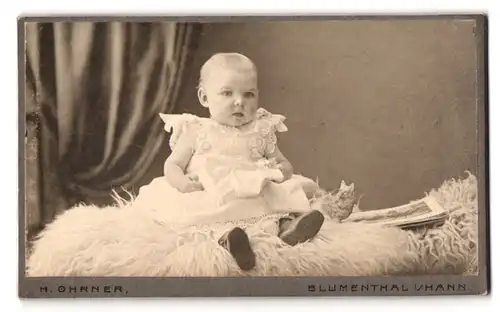 Fotografie H. Ohrner, Blumenthal i. Hann., Portrait kleines blondes Mädchen im weissen Kleid auf einem Fell sitzend