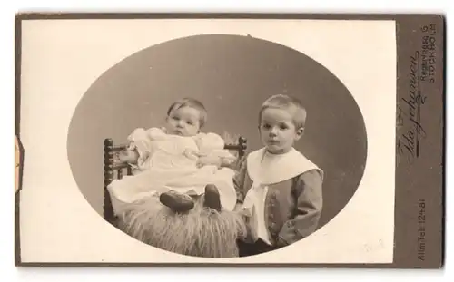 Fotografie Ida Johanson, Stockholm, Regerinsg. 6, Portrait Geschwister im Anzug mund weissen Kleid auf Fell sitzend