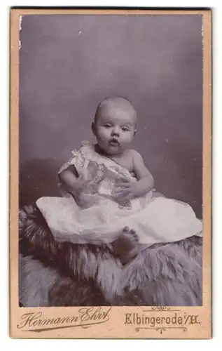 Fotografie Hermann Ehrt, Elbingerode i. H., Portrait kleines Kind im weissen Kleid sitzt auf einem Fell