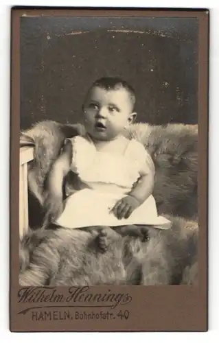 Fotografie Wilhelm Hennings, Hameln, Portrait niedliches Baby im weissen Kleidchen auf Felldecke sitzend