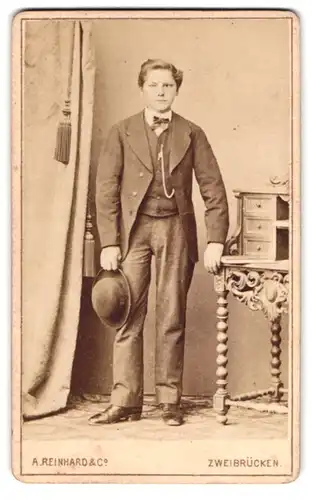 Fotografie A. Reinhard & Co., Zweibrücken, Junger Bursche in Anzug lehnt an Tisch und hält seine Melone
