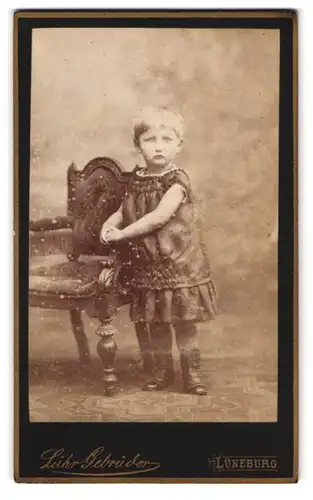 Fotografie Lühr-Gebrüder, Lüneburg, Kleines Mädchen in dunklem Kleid steht neben gepolstertem Stuhl