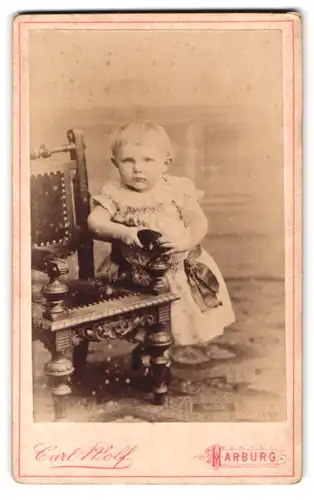 Fotografie Carl Wolf, Harburg, Kleinkind in weissem Kleidchen mit Rüschenverzierung lehnt an Stuhl