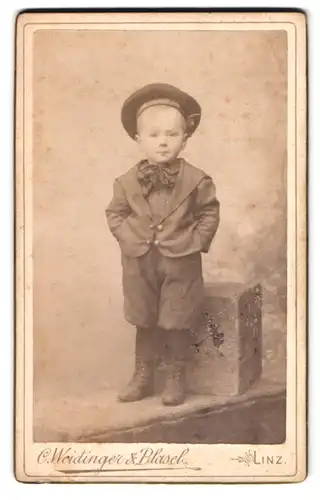 Fotografie C. Weidinger & Blasel, Linz, Kleiner Junge posiert mit Mütze und Stiefelchen