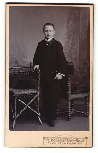 Fotografie H. Böbers, Berlin-Rixdorf, Portrait halbwüchsiger Knabe im Anzug an Stuhl gelehnt