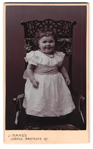 Fotografie J. Maass, Lübeck, Breitestrasse 37, glucksendes Kleinkind mit weissem Kleid auf verziertem Stuhl