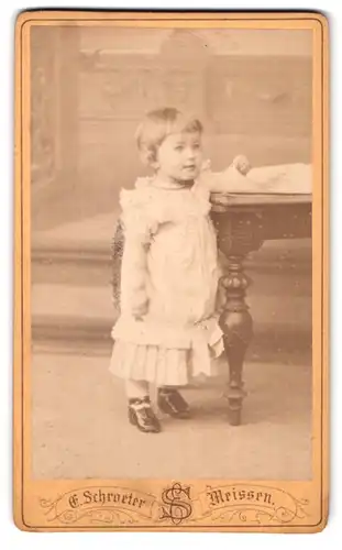 Fotografie E. Schroetere, Meissen, Obergasse 597, Portrait kleines Mädchen im weissen Kleid steht am Tisch