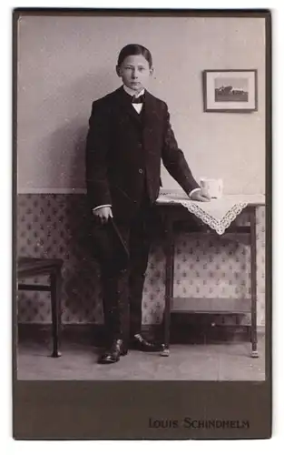 Fotografie Louis Schindhelm, Ebersbach i. S., Portrait junger Knabe im Anzug mit Hut vor einer Studiokulisse