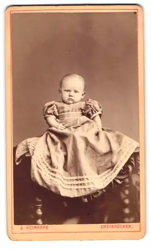 Fotografie A. Reinhard, Zweibrücken, nahe dem Bahnhof, Portrait Kleinkind im zu grossen Kleid sitzt im Atelier