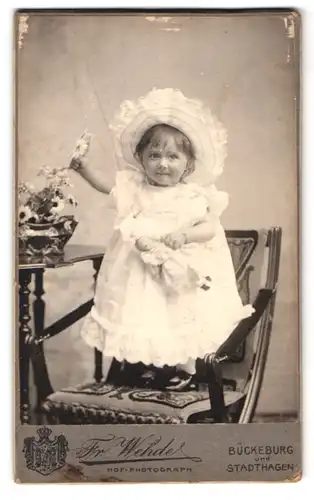 Fotografie Fr. Wehde, Bückeburg, Portrait kleines Mädchen im weissen Kleid mit Puppe und Sommerhut