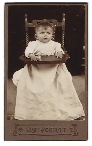 Fotografie Fotograf und Ort unbekannt, Portrait Kleinkind im zu grossen Kleid sitzt im Kinderstuhl