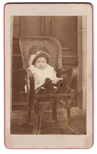Fotografie Fotograf und Ort unbekannt, Portrait kleines Kind im gestreiften Kleid mit Mütze auf einem Stuhl sitzend