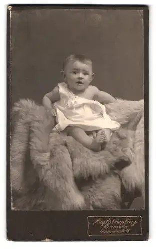 Fotografie Aug. Striepling, Hameln, Emmernstr. 18, Portrait niedliches Kleinkind im Kleid auf Fell sitzend