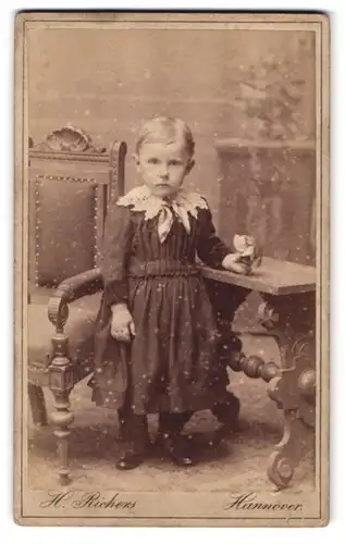 Fotografie H. Richers, Hannover, Cellerstr. 146, Portrait kleines Mädchen im Kleid mit Spitzenkragen vor Kindermöbeln
