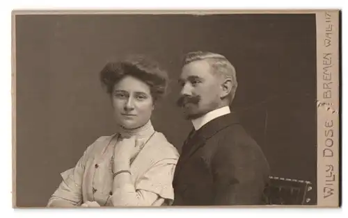 Fotografie Willy Dose, Bremen, Wall 117, Portrait Mann und Frau im Anzug mit Walrossbart und Biedermeierkleid mit Kette