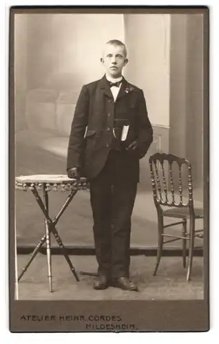 Fotografie Heinr. Cordes, Hildesheim, Zingel 36, Portrait Herr im Anzug mit Fliege hält ein Buch, Segelohren