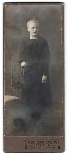 Fotografie Aug. Striepling, Hameln, Emmern-Str. 18, Portrait junges blondes Mädchen im schwarzen Kleid lehnt an Stuhl