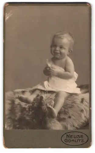 Fotografie C. Neuse, Görlitz, Blumenstr. 8, Portrait süsses lachendes Kleinkind im weissen Hemdchen auf Fell sitzend