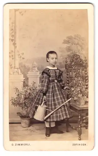 Fotografie G. Zimmerli, Zofingen, Portrait kleines Mädchen im karierten Kleid mit Gieskanne vor einer Studiokulisse