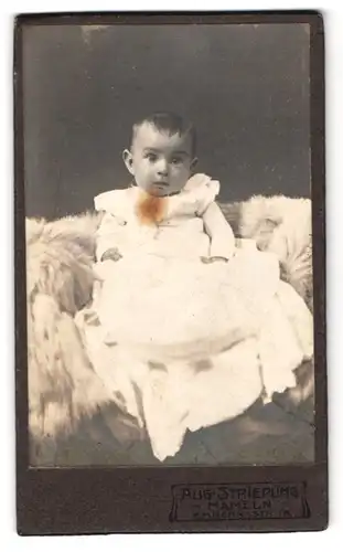 Fotografie Aug. Striepling, Hameln, Emmern-Str. 18, Portrait süsses Baby im weissen Taufkleidchen auf Fell sitzend
