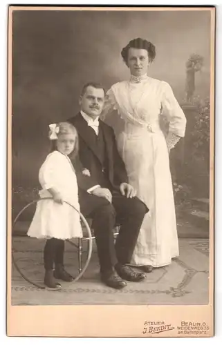 Fotografie J. Bendit, Berlin, Weidenweg 35, Mutter in weissem Kleid und sitzender Vater mit Tochter mit Spielreif