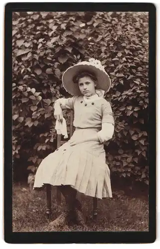 Fotografie unbekannter Fotograf und Ort, Portrait junges Mädchen im hellen Kleid mit Sommerhut sitzt im Garten