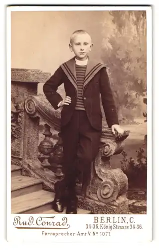 Fotografie Rud. Conrad, Berlin, König-Str. 34-36, Portrait junger Knabe im Matrosenanzug vor einer Studiokulisse