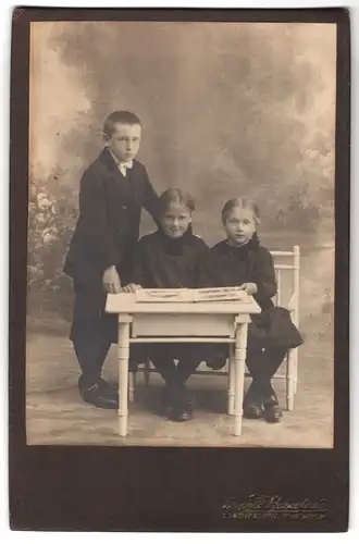 Fotografie Leop. Haberland, Eggenfelden, Stadtplatz 8, Portrait Bube steht neben einem Tisch mit zwei sitzenden Mädchen